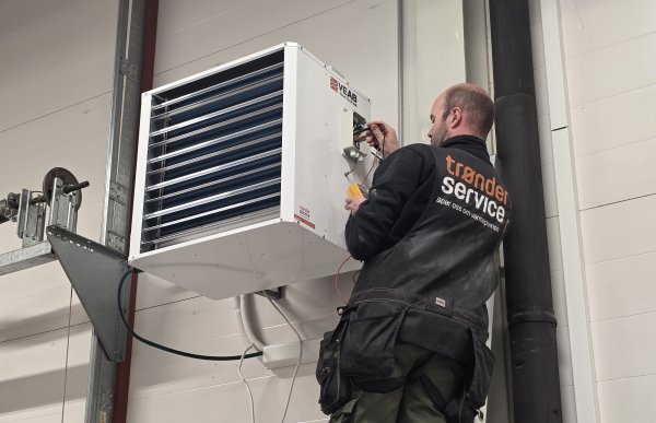 En mann i svart arbeidsjakke står med et måleinstrument som kobles til en varmepumpe på en vegg.