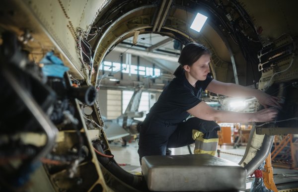 En kvinne i mørkt arbeidstøy sitter på huk inne i en stor, sirkelformet flykomponent. Hun jobber med begge hendene på ledninger og tekniske deler langs innsiden, mens arbeidslys lyser opp området.