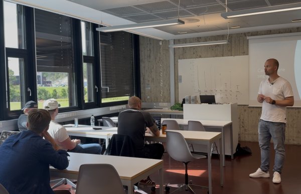 En mann står foran i et undervisningsrom og underviser en gruppe studenter som sitter ved ulike bord. Han har på seg lys T-skjorte, jeans og hvite sko. Bak ham er en tavle og et lerret, og langs den ene siden av rommet er det store vinduer.