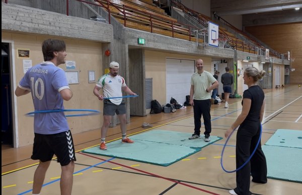 En mann står sammen med tre andre personer i en idrettshall. To menn og en kvinne bruker rockeringer, mens mannen står mellom dem og følger med. I bakgrunnen er det basketballkurver, tribune og flere andre personer.