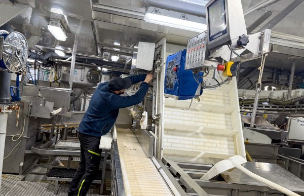 En mann med blå jakke og svarte bukser arbeider ved en maskin om bord i en tråler, omgitt av et samlebånd, metallkonstruksjoner og annet produksjonsutstyr.
