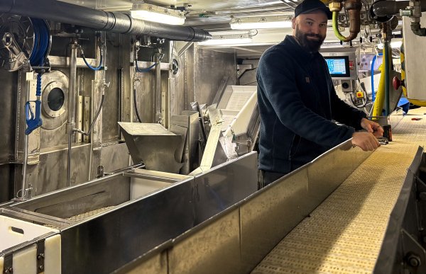 En mann med skjegg og caps arbeider ved et samlebånd. Rundt samlebåndet er det maskiner, beholdere og annet produksjonsutstyr.