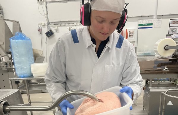 En dame står i hvitt arbeidstøy og har på seg hørselsvern. Hun holder en beholder som fylles med kjøttfarse. Hun befinner seg i et matproduksjonslokale.