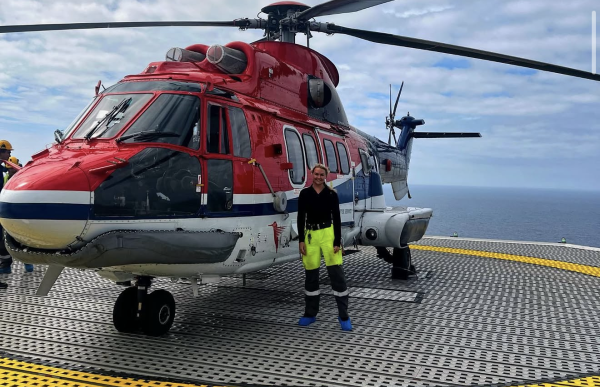 En kvinnelig arbeider står på et helikopterdekk på en offshoreplattform foran et stort rødt, hvitt og blått helikopter. Hun har på seg svart overdel og gule og svarte arbeidsbukser med refleks. Helikopteret fyller store deler av bildet, og i bakgrunnen ses åpent hav og en overskyet himmel.