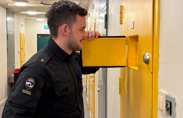 En mann med brunt hår og skjegg ser inn i ei luke i ei gul dør