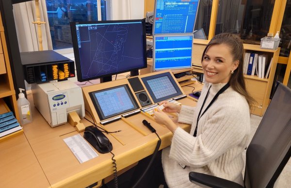 Dame med brunt hår, hvit genser og et nøkkelkort rundt halsen smiler inn i kamera. Hun sitter på et kontor med flere skjermer, både pc-skjermer og mindre skjermer. En telefon og små briller i ulike farger ligger på pulten.