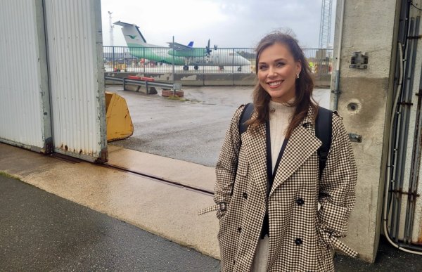 Dame med brunt hår, hvit genser og rutete jakke står foran en port med et fly i bakgrunnen.