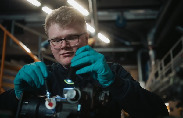 En mann med briller og turkise engangshansker bøyer seg over en mørk, sylindrisk komponent. Han holder et smalt verktøy i den ene hånden og undersøker delen på nært hold.