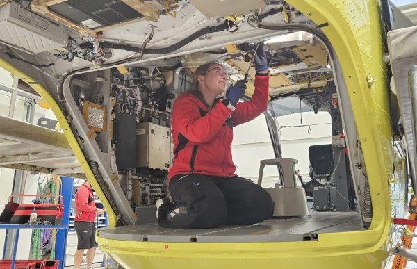 En kvinne med briller, rød arbeidsjakke og blå hansker sitter på kne inne i et gult helikopter. Hun holder et verktøy mot en komponent i taket. Deksler er fjernet, slik at ledninger, rør og teknisk utstyr er synlig rundt henne.