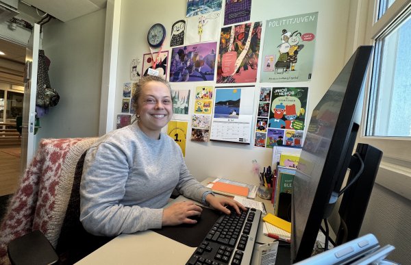 Dame sitter på et kontor foran en pc. På veggen henger det plakater av bokcover.