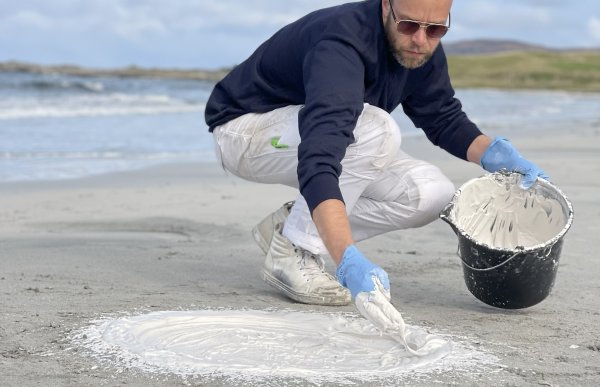 En mann sitter på huk og smører ut hvit masse i en ring. Dette foregår på en sandstrand. Han har en bøtte med hvit masse i den andre hånden. 