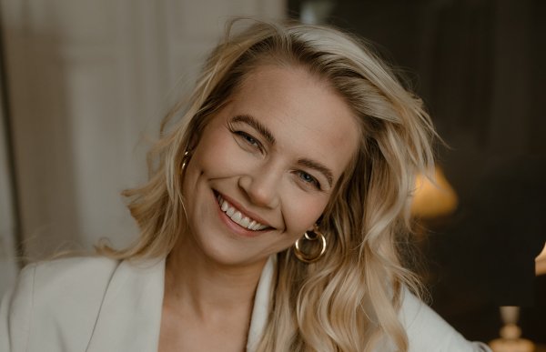Kvinne med langt lys hår smiler til kamera. Hun har på seg hvit blazer og beige topp. 