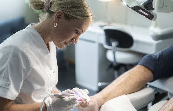 En kvinne i hvit skjorte og plasthansker og et lite stålredskap jobber med tær på en person.