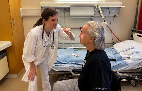 En kvinne i mørkt hår og hvit frakk holder et lite instrument med lys mot øynene til en mann i svart genser