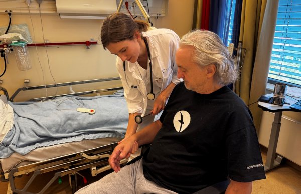 En kvinne med mørkt hår i hvit frakk holder et lite instrument på hånda til en mann i svart t-skjorte som sitter i en stol 