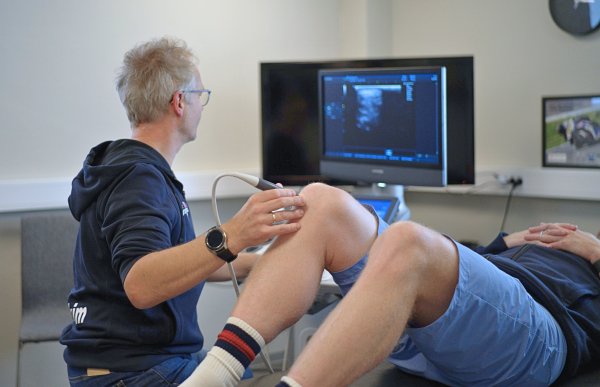 Mann i blå hettegenser. Han bruker ultralyd fra en ultralydmaskin til å studere et kne. På benken ligger en person i blå klær.