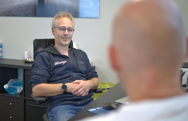 Mann i blå hettegenser lener seg tilbake i en kontorstol mens han snakker med en mann.