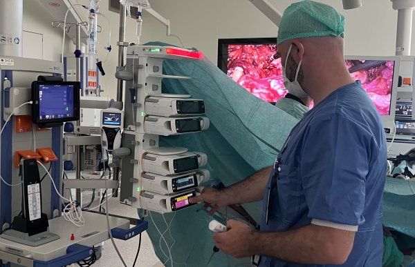 En mann står med ryggen til og hånda trykkende på et medisinsk apparat med en skjerm. Han har på seg blå medisinsk overdel, munnbind og blå hette. Rundt i rommet er det ulik medisinsk utstyr, og bak han foregår det en operasjon.