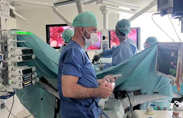 En mann i blå medisinsk skjorte, blå hette på hodet og munnbind står og ser på en skjerm hvor det står puls og andre medisinske beskrivelser av en pasient i narkose. Han holder utstyr i hånda, og rundt han er det ulik medisinsk utstyr. Bak han foregår en operasjon.