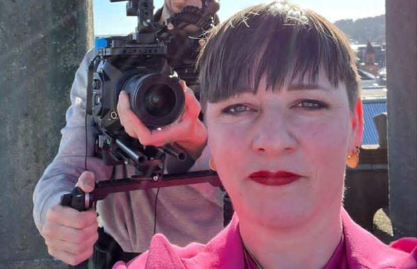 En kvinne tar en selfie i solskinnet, mens en kameramann bak henne filmer med stort videokamera. Hun har på seg rosa blazer og rød leppestift, med himmelen og bylandskap i bakgrunnen.