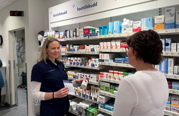 En kvinne i apotekuniform står foran hyller med kosttilskudd og snakker med en kunde. Hun holder et produkt i hånden og viser samtidig til informasjonsmateriell. Hyllene bak er fylt med ulike kosttilskudd og produkter for fordøyelse.