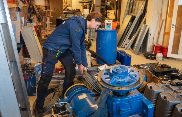 En heismontør arbeider i et verksted. Han bøyer seg over en stor blå heismotor og holder i flere ståltau som er viklet rundt motorens trommel. Rundt ham er det hyller, verktøy, reservedeler, pappesker og tekniske maskindeler.