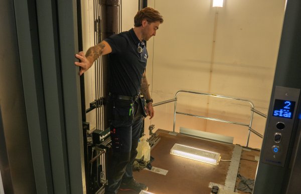 En heismontør står oppå en heis, inne i en åpen heissjakt. Han holder hånden på heisdøren mens han står på toppen av heiskabinen. Rundt ham ses stålkabler, føringsskinner og tekniske komponenter. På gulvet av kabintaket ligger to arbeidslamper som lyser opp området.