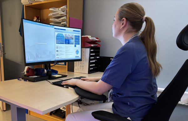 En kvinne med brunt hår i hestehale og med blå skjorte sitter foran en dataskjerm