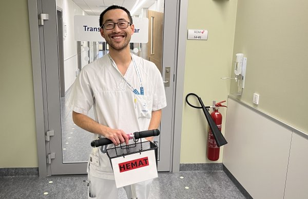 En mann i hvit sykehusklaff står smilende i en sykehuskorridor og holder en liten sparkesykkel med et skilt der det står «HEMAT». Bak ham er en glassdør inn til transfusjonsavdelingen.