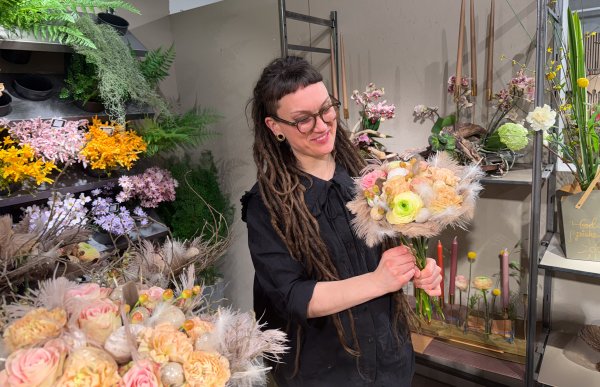Kvinne med langt brunt hår, iført svart skjorte og briller, står i butikken og holder en blomsterbukett. I bakgrunnen sees miljø fra en blomsterbutikk.