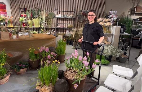 Kvinne med langt brunt hår, iført svart skjorte og briller, står foran en tralle med blomster. I bakgrunnen ser man en typisk blomsterbutikk.