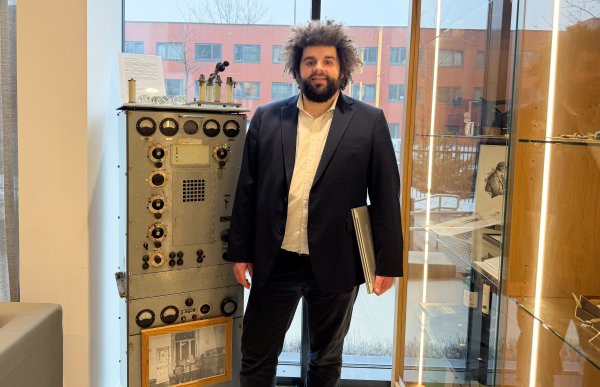 En mann med langt brunt krøllete hår, kledd i hvit skjorte, mørkeblå jakke og svarte bukser, står foran en eldre radio og poserer.