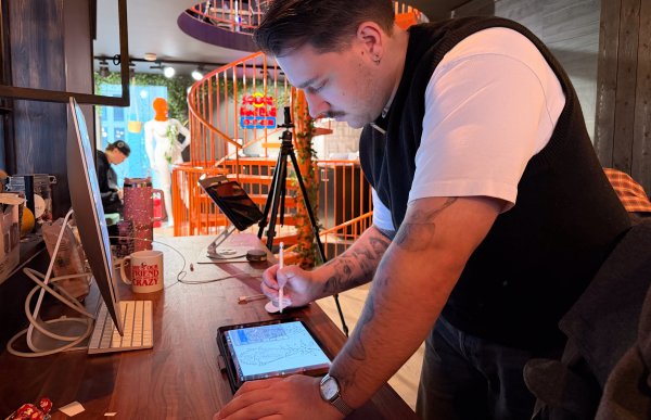 Mann med mørkt hår og hvit T-skjorte står ved et skrivebord og tegner ferdig et tatoveringsdesign på en iPad.