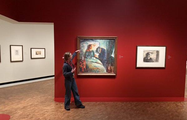 En dame i mørke klær peker på et Munch maleri som henger på veggen, hun befinner seg i et kunstmuseeum. Bakgrunnen på veggen er rød og bak denne veggen, til venstre, ser man en grå vegg med flere malerier på.