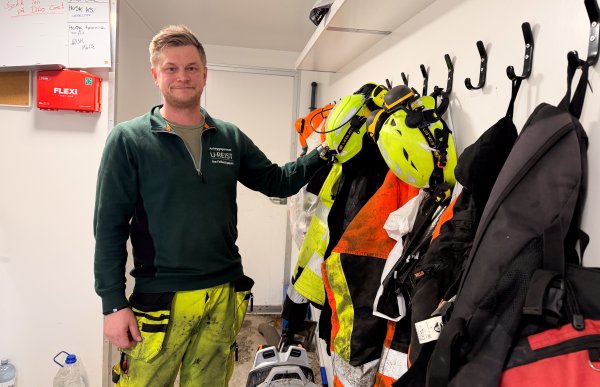 Mann med mørkeblondt hår, neon arbeidsbukse og grønn genser står inne i en brakke på anleggsplassen og poserer i et typisk brakkemiljø.