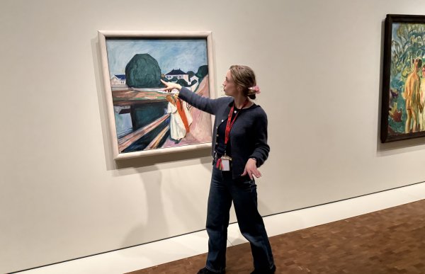 En dame i mørke klær peker på et Munch maleri som henger på veggen, hun befinner seg i et kunstmuseeum.