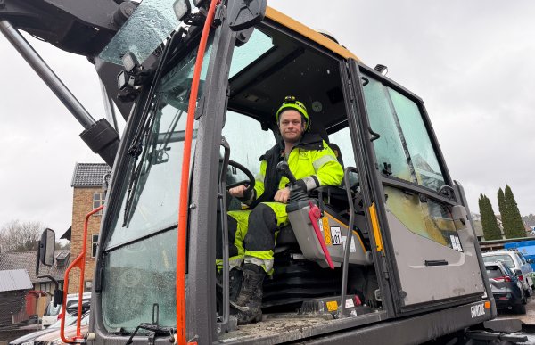 nleggsmaskinfører i neon arbeidsklær og hjelm sitter i førerhuset på en gravemaskin.
