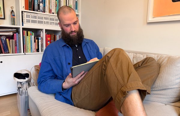 Mann med kort hår og langt mørkt skjegg, iført lyseblå jakke, mørkeblå skjorte og brun bukse, sitter i en sofa med bena delvis i kryss og skisserer på en iPad i fanget.