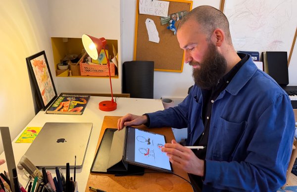 Mann med kort hår og langt mørkt skjegg, iført lyseblå jakke og mørkeblå skjorte, sitter ved pulten på kontoret og skisserer. I bakgrunnen sees blyanter og bøker. Bildet er tatt fra siden.