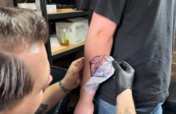 Mann med mørkt hår har nettopp plassert et tatoveringsdesign på armen til en kunde og trekker forsiktig av designpapiret