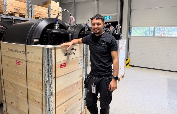 Mann med kort svart hår, svart arbeids T-skjorte og vernebukse står ved ferdigstilte produkter som er klargjort for utsending til kunder.