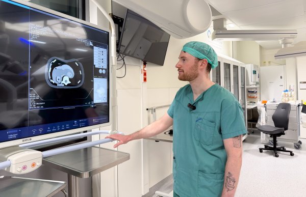Mann med oransje skjegg iført grønt kirurgisk arbeidstøy og kirurgisk hette står og ser på en stor skjerm som viser røntgenbilder. I bakgrunnen ses en operasjonssal med medisinsk teknologi og utstyr.