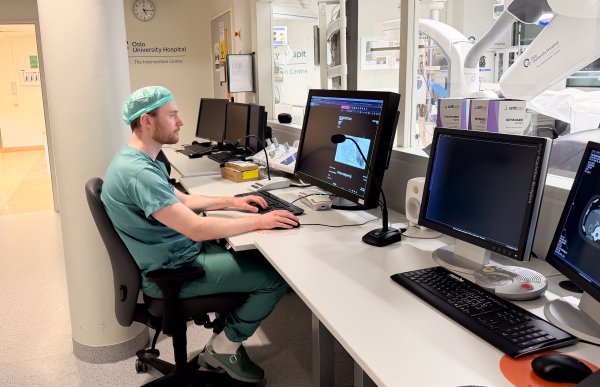 Mann med oransje skjegg iført grønt kirurgisk arbeidstøy og kirurgisk hette sitter og observerer RTG-bilder på PC-skjermer. Rundt ham er det flere PC-er i et arbeidsmiljø typisk for radiografer.