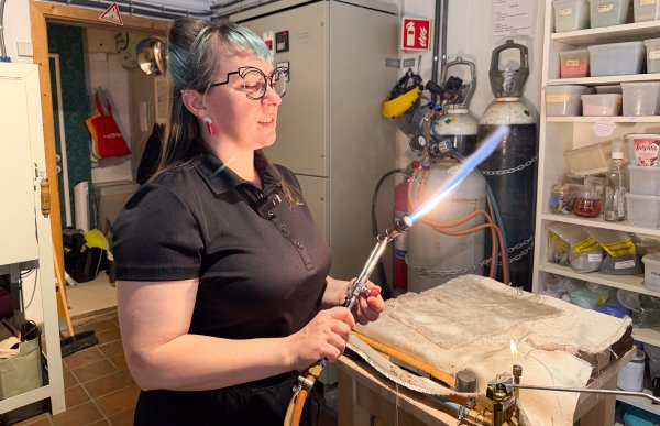 Kvinne med langt mørkt hår og turkis pannelugg, iført briller og svart T-skjorte, holder en gassbrenner med synlig flamme. Bildet viser glassblåseren i arbeid, og i bakgrunnen sees utstyr og arbeidsmiljø fra verkstedet.