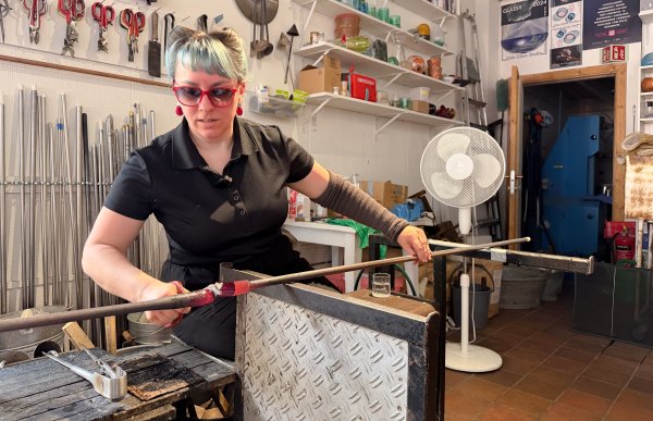 Glassblåser med langt mørkt hår og turkis pannelugg holder en lang metallstang med glødende, rødt glass på enden. Hun bruker et verktøy til å forme og klippe glasset. I bakgrunnen sees utstyr og arbeidsmiljø fra glassblåserverkstedet.