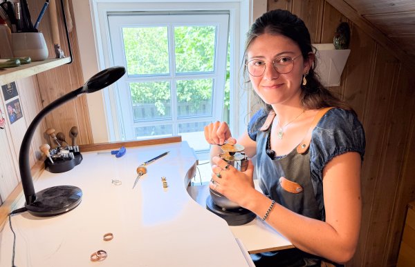 Kvinne med langt brunt krøllete hår sitter ved arbeidsstasjonen sin iført arbeidsforkle og arbeider med gravering. Hun holder et graveringsverktøy og ser mot kamera.