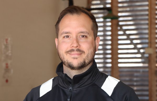 En mann i svarte og gule arbeidsklær smiler mot kamera. Han har mørkt, kort hår og bart og skjegg.