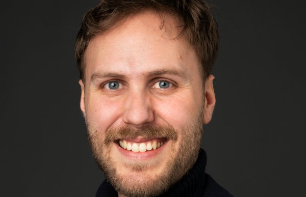 Mann med mørkebrunt hår, skjegg og bart smiler bredt og ser rett inn i kameraet. Han er kledd i en svart turtleneck-genser og en svart dressjakke. Bakgrunnen er en ensfarget, mørkegrå studiofarge.