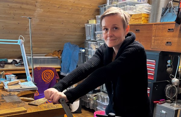 En kvinne med kort, lyst hår. Hun er kledd i en svart genser og rosa arbeidsbukser. Kvinnen holder i en metallflatningsmaskin. Hun er i et verksted.