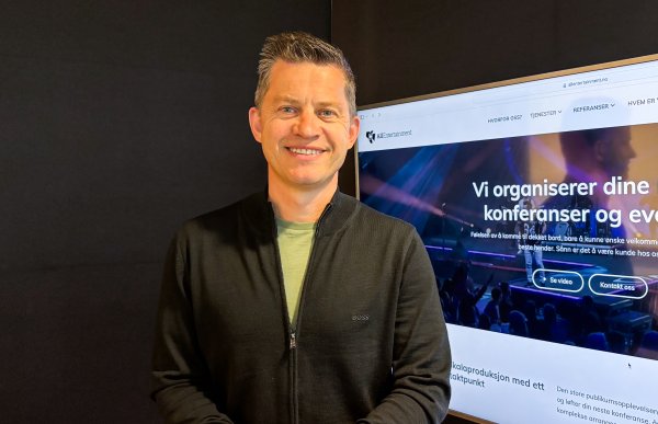 Mann med kort, mørkt hår smiler mot kamera. Han har på seg svart genser med en lys t-skjorte synlig i åpningen. En TV står på i bakgrunnen, inne på kontoret hans.